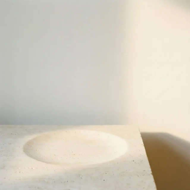 A minimalist scene showing a light-colored, textured stone or concrete surface with a shallow circular indentation, softly lit by natural sunlight casting gentle shadows. The background is a plain, light wall. {{brizy_dc_image_alt imageSrc=