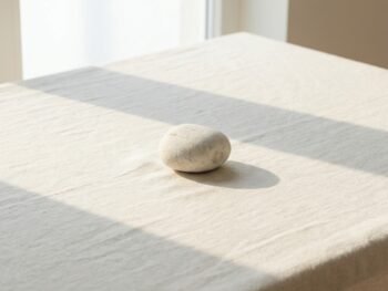 A smooth, round stone rests on a light-colored, textured cloth on a table, with soft sunlight streaming in through a nearby window and casting gentle shadows. {{brizy_dc_image_alt entityId=